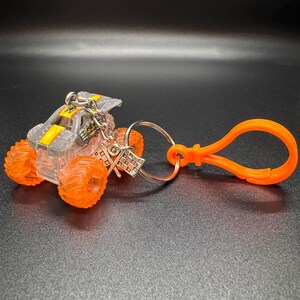 May include: A miniature monster truck keychain. The truck has a clear body with gray and yellow accents, and bright orange wheels. It is attached to a silver chain and an orange clip.