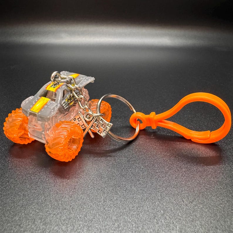 May include: A miniature monster truck keychain. The truck has a clear body with gray and yellow accents, and bright orange wheels. It is attached to a silver chain and an orange clip.