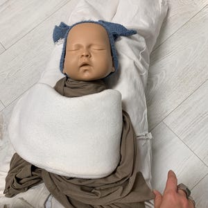 May include: A white fabric swaddle with a brown fabric wrap and a light blue knitted hat on a baby doll. The doll has a light brown complexion and closed eyes.