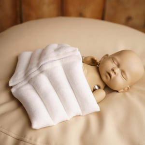 May include: A white, rectangular, quilted fabric heating pad with a visible zip closure, positioned over the torso of a light-skinned, bald doll with closed eyes, resting on a smooth, beige, rounded surface.