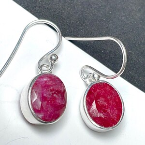 May include: A pair of silver dangle earrings featuring oval-cut, deep red gemstones. The gemstones are set in a bezel setting, and the earrings have a simple hook closure. The earrings are displayed against a white and black background.