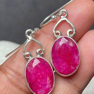 Könnte beinhalten: Silberne Ohrringe mit oval geschliffenen, leuchtend rosa Edelsteinen. Jeder Edelstein ist in einen silbernen Rahmen mit einem dekorativen Wirbelmuster gefasst. Die Ohrringe haben einen Hakenverschluss.