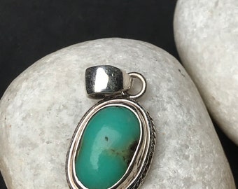May include: A silver pendant with an oval, light-green gemstone. The stone is set in a silver frame with a braided edge. The pendant has a small loop at the top for a chain. The background is a white stone against a black backdrop.