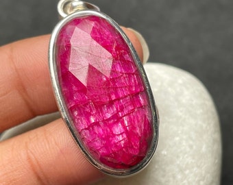 May include: A faceted, oval-shaped ruby pendant with a silver-colored bezel. The ruby is a vibrant shade of pink-red, with visible internal textures and light reflections. The pendant has a small loop at the top for a chain.