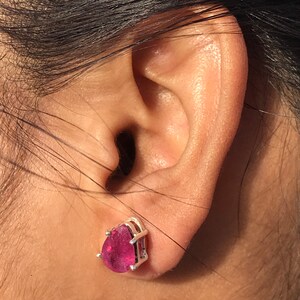 May include: A close-up of a pink teardrop-shaped gemstone stud earring set in a silver metal frame. The earring is worn on an ear, with dark hair visible in the background. The gemstone has a vibrant, saturated color.