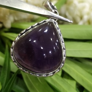 May include: A silver pendant with a dark purple amethyst gemstone. The pendant is in the shape of a teardrop and has a detailed silver border.