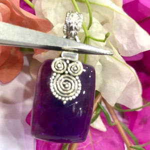 May include: A rectangular, deep purple pendant with a silver-colored bail and decorative swirls. The pendant is held by tweezers against a backdrop of pink and white flowers. The silver bail has a detailed design.