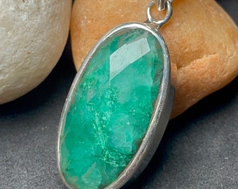 May include: A silver pendant with a large, faceted emerald gemstone. The emerald is a vivid green and set in a polished silver bezel. The pendant has a simple, elegant design, suitable for everyday wear.