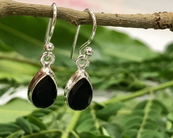 May include: A pair of silver drop earrings with black teardrop-shaped gemstones. The earrings are hanging from a small branch against a blurred green and pink background. The gemstones are faceted, catching the light.