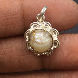 May include: A silver pendant with a floral design surrounding a cream-colored, iridescent stone. The pendant has a small loop at the top for a chain. The stone has a subtle, textured appearance.