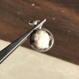 May include: A small, off-white pendant with a carved design, held by silver tweezers. The pendant is encased in a silver circular frame with cut-out details. The background is a blurred wooden surface.