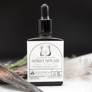 May include: A black glass bottle of "Spirit Speak Magic Ritual Oil" with a dropper, a white label, and text. The label includes the words "Sweet Tobacco, Oak Barrel, & Embers." The bottle is 1 fl oz. A feather and wood are in the foreground.