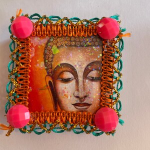May include: A square framed image of a Buddha with a green, orange, and gold trim. The frame is decorated with pink, faceted beads.
