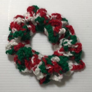 May include: A circular hair scrunchie made of crocheted yarn in a variegated pattern of red, white, and dark green. The accessory features a ruffled, textured appearance with a central opening, constructed from soft, intertwined fiber loops.