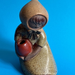 May include: A ceramic figurine of a girl wearing a brown hooded cloak and holding a red pot. The figurine has a rustic, hand-painted look.