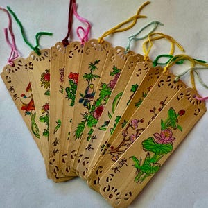May include: A collection of wooden bookmarks with colorful floral and bird designs. Each bookmark features a unique hand-painted illustration and decorative cut-out edges. The bookmarks have colorful string tassels in various colors.