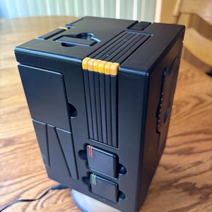 May include: A black, rectangular electronic device with a yellow and black design. The device has two buttons with red and green accents. The device is sitting on a wooden surface.