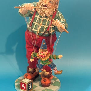 May include: A figurine of Santa Claus operating a puppet. Santa wears a green plaid shirt, red pants, and suspenders. The puppet is a clown with a blue hat and red and white striped legs. A block with the letters "AB" is on the base.