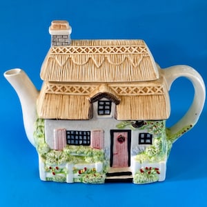 May include: A ceramic teapot shaped like a cottage with a thatched roof, chimney, and a door. The teapot has a white fence, pink shutters, and green foliage. The handle and spout are white, and the background is blue.