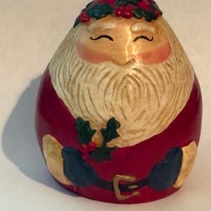May include: A painted Santa Claus figurine with a cream-coloured beard and face, red suit, and a green and red holly wreath. The figurine is egg-shaped and has a serene expression.