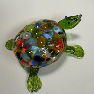 May include: A small glass turtle figurine with a multi-colored shell. The shell is made of small, round pieces of glass in various colors, including red, green, blue, yellow, and orange. The turtle has a green body and head, and its legs are also green.