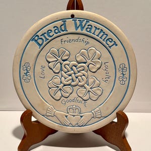 Puede incluir: Un calentador de pan redondo de cerámica color hueso con texto azul que dice Bread Warmer, Friendship, Love, Loyalty y Goodluck. El centro muestra un diseño de nudo en relieve, tréboles de cuatro hojas y un símbolo Claddagh.