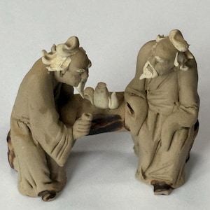 Op de afbeelding: Een keramische sculptuur met twee oudere figuren die aan een tafel zitten. De figuren hebben lange baarden en traditionele kleding. Op de tafel staan kleine keramische objecten. De sculptuur is in tinten van beige en bruin.