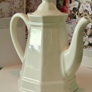 May include: A cream-colored ceramic teapot with a hexagonal body, a matching lid with a pointed finial, a curved handle, and a tapered spout. The teapot features a smooth, glossy finish and a structured, geometric design with defined vertical edges.