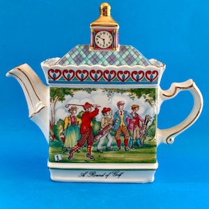 Könnte beinhalten: Dekorative Porzellan-Teekanne mit Golf-Motiv. Die Teekanne hat einen Uhrturm auf der Oberseite und eine gemalte Szene von Golfspielern. Auf der Unterseite der Teekanne steht "A Round of Golf". Die Teekanne hat einen weißen Hintergrund mit goldenen Verzierungen.