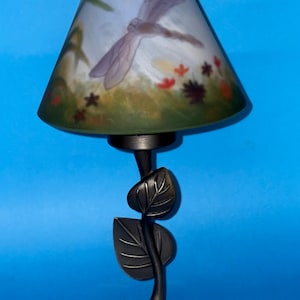 Może przedstawiać: Dekoracyjna lampa z motywem ważki na kloszu. Klosz ma kształt stożka z namalowaną ważką, kwiatami i trawą. Podstawa ma wzór liści i ciemnobrązowe wykończenie.