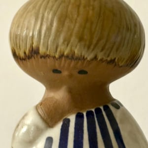 Może przedstawiać: Ceramiczna figurka przedstawiająca stylizowaną głowę z gładką, jasnobrązową, zaokrągloną fryzurą i dwiema małymi ciemnymi kropkami zamiast oczu. Postać ma na sobie biały ubiór ozdobiony pionowymi i poziomymi ciemnoniebieskimi paskami.