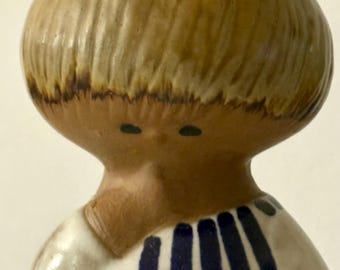 May include: A ceramic figurine featuring a stylized head with a smooth, light brown, rounded hairstyle and two small dark dots for eyes. The figure wears a white garment decorated with vertical and horizontal dark blue stripes.