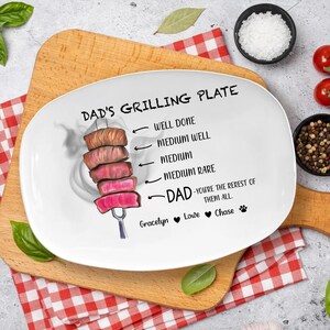 Könnte beinhalten: Weiße ovale Grillplatte mit dem Text "DAD'S GRILLING PLATE" und einer Grafik von gegrillten Steakstücken. Die Platte enthält auch den Text "WELL DONE" bis "MEDIUM RARE" und "DAD - YOU'RE THE REREST OF THEM ALL."