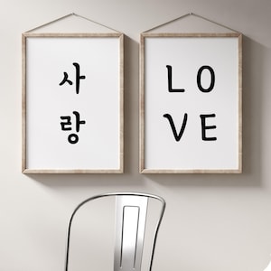 Puede incluir: Dos impresiones enmarcadas en una pared blanca. La impresión de la izquierda dice "사랑" en coreano. La impresión de la derecha dice "LOVE" en inglés.