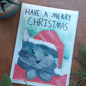 May include: A Christmas card featuring a watercolor illustration of a gray cat wearing a red Santa hat. The card has the text "HAVE A MERRY CHRISTMAS" written on it.