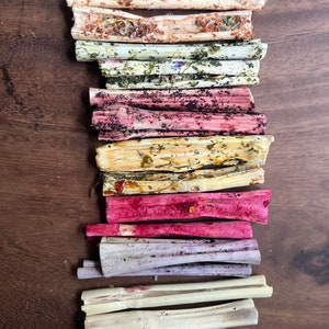 May include: A stack of 10 natural, dried, and dyed sticks. The sticks are various shades of pink, purple, yellow, and brown. The sticks are arranged vertically, with the top stick being the lightest color and the bottom stick being the darkest color.