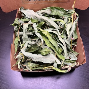 Puede incluir: Una caja de cartón con una ventana de plástico transparente, llena de hojas verdes secas. La caja está sobre una superficie oscura.