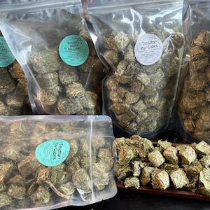 May include: Five clear plastic bags filled with Timothy Hay Cubes. The bags are labeled "Timothy Hay Cubes". There is a small pile of Timothy Hay Cubes on a wooden surface.