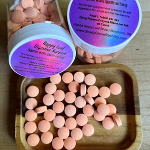 May include: Orange, round tablets are scattered on a wooden tray and inside two clear plastic containers. The containers have white lids with purple labels that read "Happy Loaf Digestive Support." The text on the label includes "Digestion, Motility, Appetite and Energy."