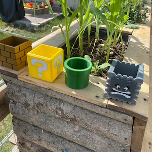 Könnte beinhalten: Vier unterschiedlich geformte Pflanztöpfe, die wie Gegenstände aus dem Videospiel Super Mario Bros. aussehen. Die Töpfe sind aus Kunststoff und in verschiedenen Farben bemalt, darunter Braun, Gelb, Grün und Grau. Die Töpfe stehen auf einer Holzoberfläche.