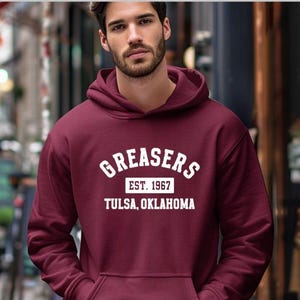 May include: Burgundy hoodie with the word "GREASERS" in white block letters, with "EST. 1967" and "TULSA, OKLAHOMA" below. The hoodie has a drawstring hood and a front pocket.