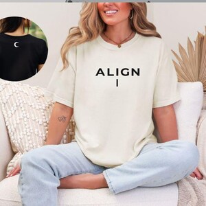 May include: Cream-colored t-shirt with the word "ALIGN" in black capital letters, with a vertical line below. The image also shows a black t-shirt with a white crescent moon on the back. The person is wearing light blue jeans.