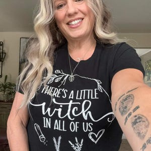 May include: A black short-sleeved t-shirt featuring white text that reads There's a little witch in all of us. The design includes a white line-art illustration of a salt shaker, sprigs of herbs, a heart, and a decorative leafy border.