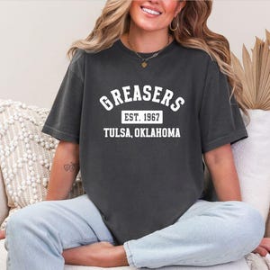 May include: Dark gray t-shirt with the word "GREASERS" in white arched lettering, "EST. 1967" in a rectangle, and "TULSA, OKLAHOMA" below. The shirt is worn by a person sitting on a white chair.