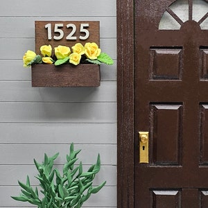 May include: A wooden house number sign with the number 1525 on it. The sign is mounted on a gray wall with a brown door to the right. There are yellow flowers in a planter box below the sign.