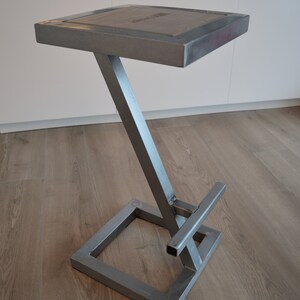 Pu&ograve; includere: Uno sgabello da bar in metallo argentato con un piano in legno. Lo sgabello ha una base a forma di Z e un poggiapiedi quadrato.
