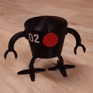 Puede incluir: Figura de robot negro con cabeza cilíndrica, brazos y piernas. El robot tiene un círculo rojo en la parte delantera y el número "02" en blanco. Los brazos terminan en forma de garra.