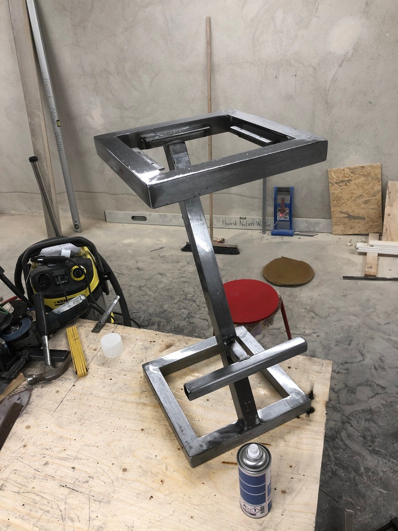 Pu&ograve; includere: Uno sgabello in metallo con una seduta quadrata e una base a forma di X unica. Lo sgabello &egrave; dipinto di grigio e si trova su una superficie di legno.