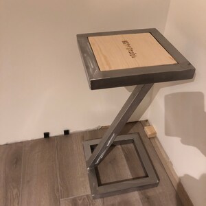 Pu&ograve; includere: Un piccolo tavolo quadrato in legno con una cornice in metallo. Il piano del tavolo &egrave; in legno chiaro e ha le parole "Iron Design" incise. La cornice metallica &egrave; argentata e ha un design semplice e moderno.