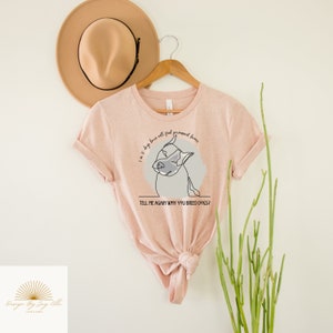 May include: A light pink t-shirt with a dog graphic and the text "I'm 10 dogs have all found permanent homes. Tell me again why you breed dogs?". A tan hat and a wooden hanger are behind the shirt.